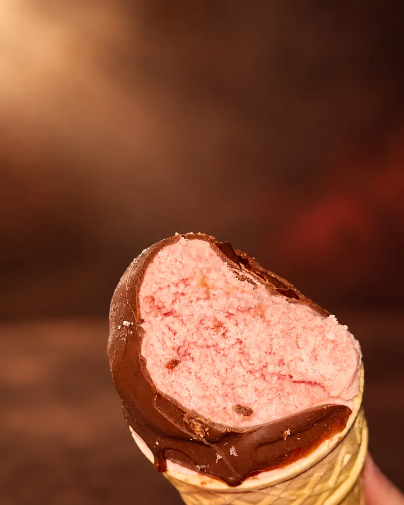 Cone Gelatto mordido, com sorvete rosa e cobertura de chocolate, fotografado em estúdio.
